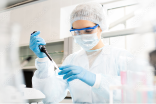 Serious concentrated female microbiologist in sterile clothing and safety goggles sitting at table and dropping reagent in petri dish while doing research in laboratory