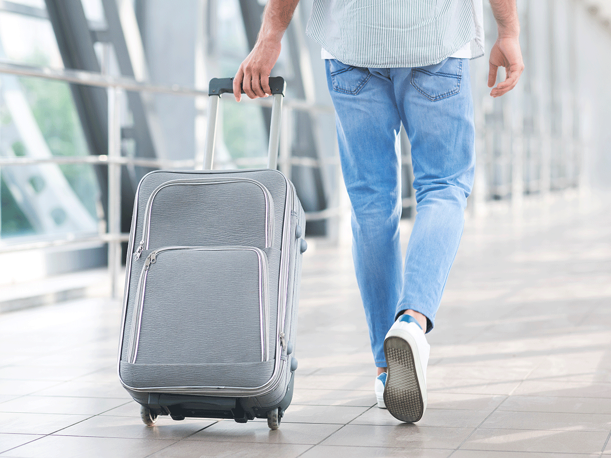 Behind view of a man walking through an airport pulling a gray wheeled suitcase